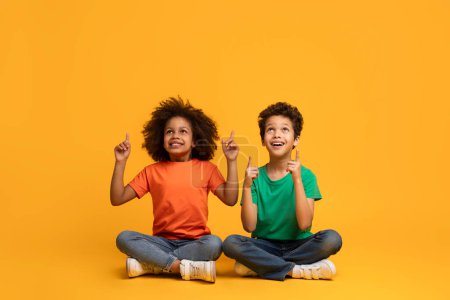 Deux enfants afro-américains, un garçon et une fille, sont assis les jambes croisées sur le sol avec les doigts tendus vers le haut, fond jaune