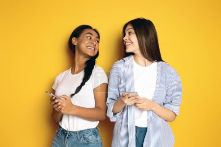 Zwei multiethnische Mädchen, eines mit Zopf und das andere mit langen Haaren, stehen nebeneinander vor einem leuchtend gelben Hintergrund und teilen einen freudigen Moment. Jeder hält ein Smartphone in der Hand