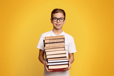 Foto de Lindo estudiante diligente en gafas con un montón de libros en las manos, fondo de estudio naranja - Imagen libre de derechos