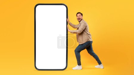 A cheerful man is standing next to a disproportionately large smartphone model with white blank screen, yellow background, mockup, copy space