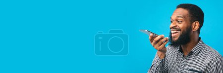Téléchargez les photos : Joyeux jeune homme noir utilisant assistant vocal sur smartphone, isolé sur un mur de studio bleu, espace de copie, bannière web - en image libre de droit