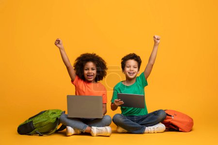 Photo for Two African American children, a boy and a girl, are sitting on the floor with laptop and digital tablet in front of them, raising hands up, yellow background - Royalty Free Image