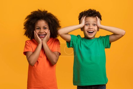African American young girl and boy are standing against a vivid yellow backdrop. Both children exhibit genuine expressions of happiness and surprise.