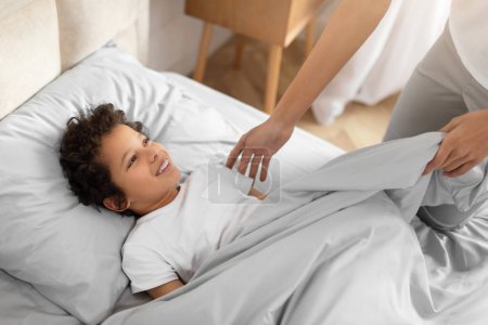 Ein fröhliches, junges afroamerikanisches Kind liegt in einem Bett, bedeckt mit einer hellgrauen Bettdecke, während ein Elternteil sie sanft in sich hineinstopft, was für ein gemütliches und sicheres Gefühl vor dem Schlafengehen sorgt.