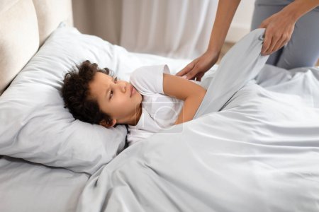 Afroamericano niño pequeño descansa en una cama con una mirada de anticipación, viendo como un padre amorosamente ajusta la manta sobre ellos