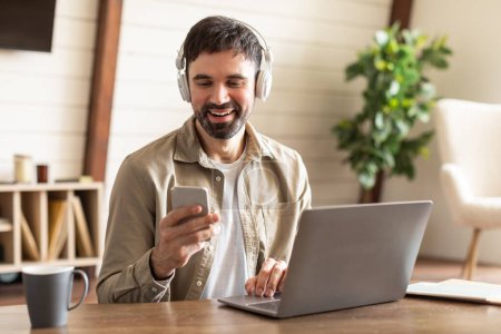 A bearded man is enjoying his time working from a home office setup, with a laptop open in front of him and headphones over his ears. He is holding a smartphone, looking at its screen with a smile