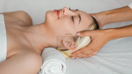 A serene atmosphere envelops a young woman as she enjoys a soothing head massage, eyes closed, melding relaxation with the ambient tranquility of a day spa setting