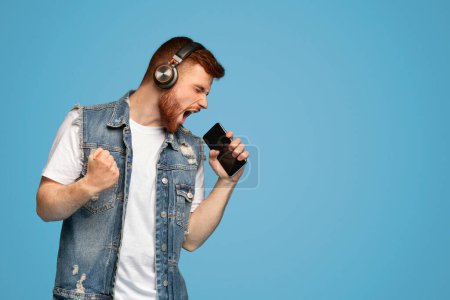 Téléchargez les photos : Jeune homme aux cheveux roux émotionnel millénaire chantant sa chanson préférée sur smartphone comme microphone sur fond bleu, espace de copie - en image libre de droit