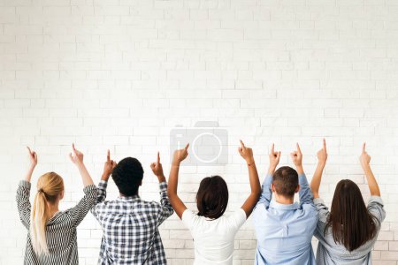Foto de Un grupo de amigos se para frente a una pared blanca en blanco, todos apuntando hacia arriba. Sus gestos sugieren que están indicando o llamando la atención sobre algo por encima de ellos, retrospectiva - Imagen libre de derechos