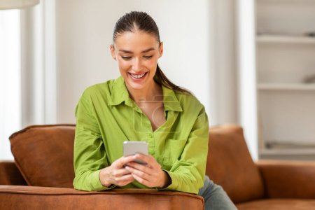 A woman is seated on a couch, engrossed in her cell phone. She appears focused as she interacts with the device, possibly texting or browsing social media.
