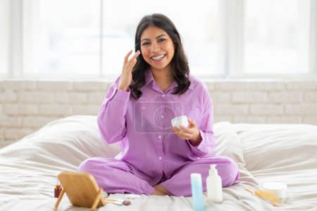 Eine strahlende junge Frau aus dem Nahen Osten sitzt im Schneidersitz auf ihrem Bett und trägt einen bequemen lila Pyjama auf, während sie sanft Feuchtigkeitscreme auf ihr Gesicht aufträgt.