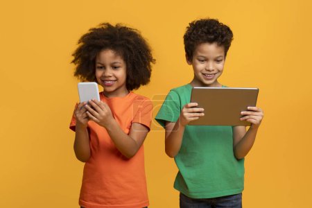 Foto de Dos jóvenes niños afroamericanos, posiblemente hermanos o amigos, están de pie uno al lado del otro sosteniendo una tableta y un teléfono celular - Imagen libre de derechos
