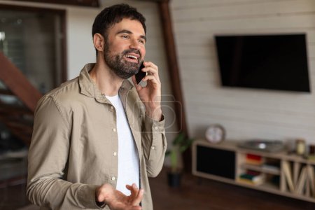 Photo for A man is engaged in a phone call while standing in a modern living room, holding a cell phone to his ear. The room is tastefully decorated with a sofa, coffee table, and plants, copy space - Royalty Free Image