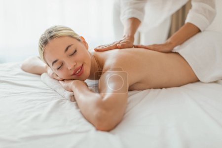 Mujer rubia está tumbada boca abajo en una mesa de masaje en un spa, mientras que un terapeuta de masaje está masajeando su espalda con presión constante.