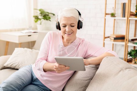 Téléchargez les photos : Femme âgée impressionnante regarder des vidéos sur tablette PC dans les écouteurs sans fil, profiter des technologies modernes, espace libre - en image libre de droit