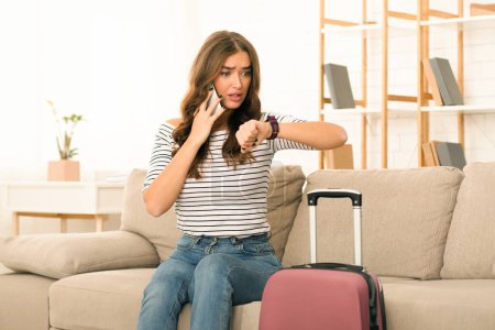 A concerned young woman sits on the edge of a couch, gazing at her watch with a look of anxiety on her face. She is preparing to depart, evident by a suitcase beside her