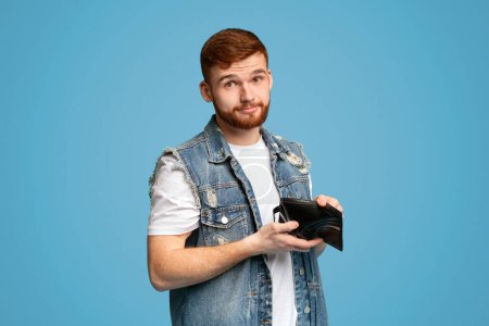 No money for living. Sad millennial man showing his empty wallet to camera on blue background, copy space