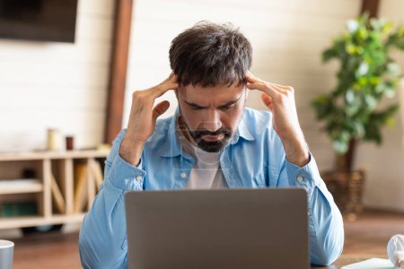Photo for A man sits at a wooden desk in a home office, showing signs of frustration while working on his laptop. He is holding his head in his hands - Royalty Free Image