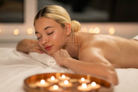 A woman is laying on top of a bed next to a row of burning candles at spa. The room is dimly lit, casting a warm glow on the scene.