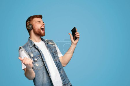 Téléchargez les photos : Cool jeune homme barbu chantant dans les écouteurs et appréciant la musique avec les yeux fermés sur fond bleu, espace de copie - en image libre de droit