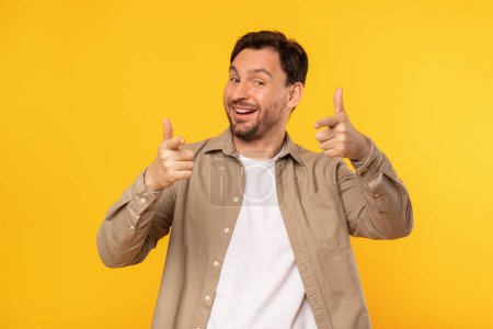 A cheerful man stands against a vivid yellow backdrop, giving a double thumbs up gesture while smiling broadly, pointing at camera