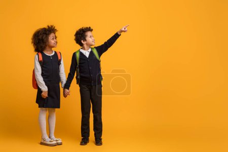 Téléchargez les photos : Un jeune garçon afro-américain et une écolière pointent vers un objet ou une direction indiscernable sur fond jaune. Leurs doigts sont tendus et ils regardent attentivement au même point. - en image libre de droit