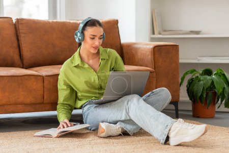 A young woman is seated on the floor of a well-lit living room with her back against a couch, focused on a laptop placed on her lap. She wears casual clothing and headphones, and has a notebook