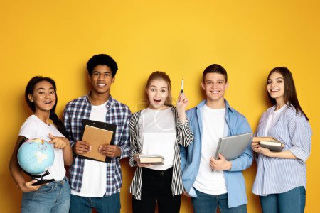 Foto de A cheerful, multiethnic group of students stands against a bright yellow backdrop, each holding different educational tools. - Imagen libre de derechos