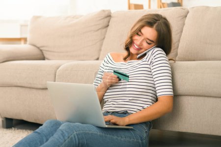 Photo for A cheerful young woman is seated on the floor leaning against a couch in a well-lit living room. She is holding a credit card and looking at her laptop screen, likely making an online purchase. - Royalty Free Image