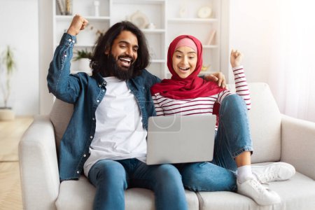 Téléchargez les photos : Un couple musulman en tenue décontractée recherche en ligne, assis ensemble dans un salon qui rayonne de chaleur et de convivialité - en image libre de droit