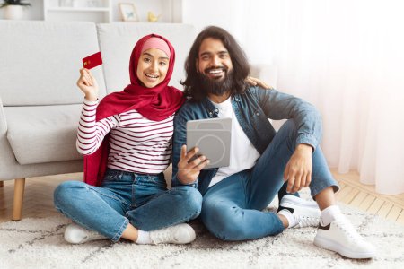 Una alegre pareja oriental se sienta casualmente en el suelo, usando una tableta digital. La mujer, con un hiyab rojo y una camisa a rayas, sostiene una tarjeta de crédito, lo que sugiere una actividad comercial o financiera