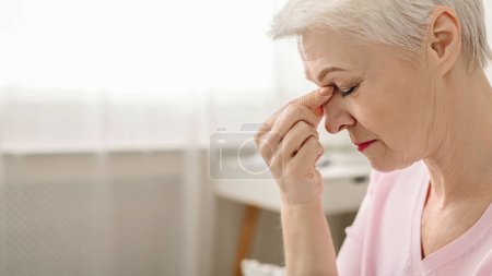 Fatigued senior woman massaging nose bridge, feeling eye strain or headache trying to relieve pain, thinking of problems, empty space