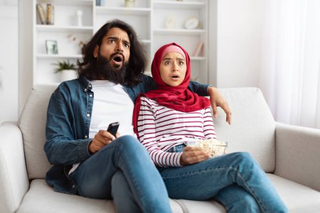 Indian man and woman are sitting side by side on a white couch in a well-lit living room. Both appear surprised and shocked by something they see on the television screen.