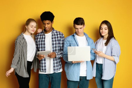 Foto de Cuatro adolescentes multiétnicos se enfrentan a un fondo amarillo brillante, cada uno profundamente centrado en su propia pieza de tecnología. - Imagen libre de derechos
