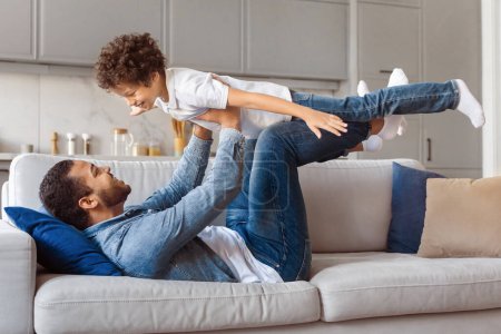 African American father lying on his back on grey sofa lifts his young son in the air with his feet, simulating an airplane flight, a moment of pure joy and bonding shared between parent and child