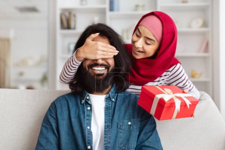 Eastern woman wearing a red hijab is surprising a bearded man by covering his eyes from behind with one hand while holding a red and white gift box with the other