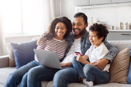 Photo for African American man, woman, and child are seated on a sofa. They are all looking at a laptop screen, engaged in an activity. The room is well-lit and cozy, creating a warm atmosphere. - Royalty Free Image