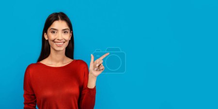 Photo for A cheerful woman with long hair wearing a red blouse stands against a vibrant blue backdrop. She has a bright smile and pointing at copy space - Royalty Free Image
