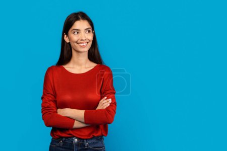 Une femme portant un pull rouge se tient debout les bras croisés dans une pose neutre. Elle semble confiante et affirmée avec son langage corporel, regardant l'espace de copie