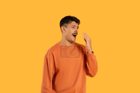 Ein junger Mann mit dunklen Haaren steht vor einem leuchtend gelben Hintergrund und bedeckt seinen Mund mit der Hand, während er weit gähnt und in einem lässig orangefarbenen Sweatshirt Anzeichen von Müdigkeit oder Langeweile zeigt..