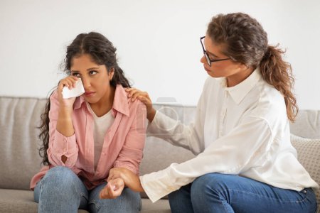 A young woman appears visibly upset, wiping her tears with a tissue as she sits on a grey sofa. Beside her, a supportive psychologist is placing a comforting hand on her shoulder