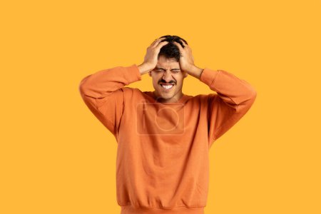 Un hombre con una camisa naranja se muestra sosteniendo su cabeza en un gesto de angustia o agotamiento. Su expresión facial transmite sentimientos de frustración o abrumamiento