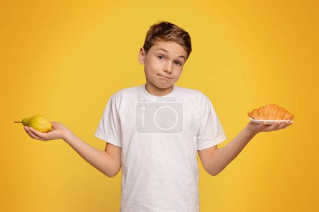 Foto de Es una decisión difícil. Adolescente pensativo elegir entre croissant y pera fresca, fondo de estudio naranja - Imagen libre de derechos