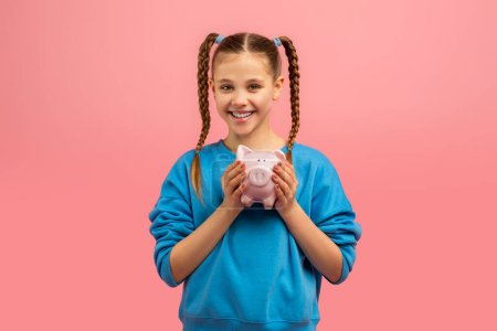 Photo for A cheerful young girl with braided hair holding a pink piggy bank and smiling against a pink background - Royalty Free Image