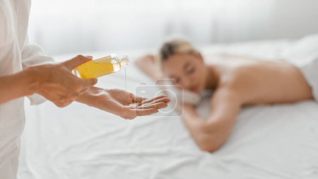 Ein professioneller Massagetherapeut wird dabei erwischt, wie er eine Behandlung vorbereitet, indem er ihnen Massageöl in die Hand gießt, während ein entspannter Kunde mit dem Gesicht nach unten im Hintergrund liegt.
