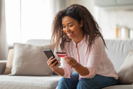 Foto de Joven mujer afroamericana alegre con tarjeta de crédito utilizando el teléfono inteligente en casa, sentado en el sofá en el interior de la sala de estar cómoda - Imagen libre de derechos