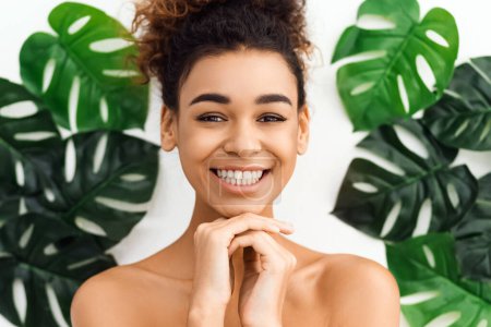 Téléchargez les photos : Femme afro-américaine souriant joyeusement avec les mains tenues près du visage dans un environnement spa, encadrée par des feuilles vertes tropicales suggérant la fraîcheur - en image libre de droit