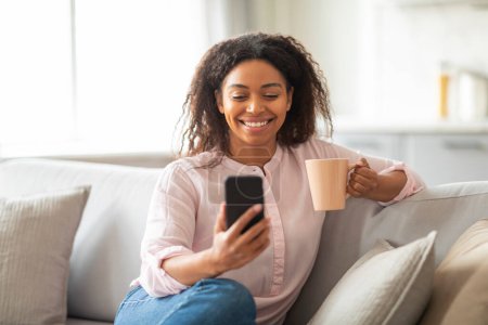 Schwarze Dame zu Hause genießt einen Kaffee, während sie durch ihr Handy blättert und verkörpert einen entspannten und modernen Lebensstil