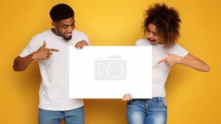 Téléchargez les photos : Jeune heureux couple afro-américain pointant vers le panneau publicitaire vierge, fond orange, espace vide pour le texte - en image libre de droit
