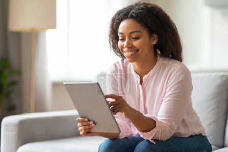 Téléchargez les photos : Une dame afro-américaine détendue sourit en naviguant sur sa tablette, soulignant l'utilisation des technologies de loisirs dans un environnement domestique - en image libre de droit